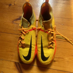 Gula Nike fotbollsskor med svart swoosh - Säljer ett par gula Nike fotbollsskor med svart swoosh och orange detaljer. Skorna har snörning och är designade för bra grepp på planen med dobbar undertill. Perfekta för dig som vill sticka ut på fotbollsplanen.