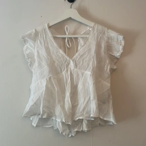 Vit blus med volang från Nelly - Vit blus från Nelly med volangdetaljer och knytning i ryggen. Blusen har v-ringning, korta ärmar och spetskant längs ärmarna. Använd 1 gång så i bra skick.