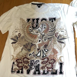 Vit t-shirt med grafiskt tryck från Just Cavalli - Säljer en vit t-shirt från Just Cavalli i storlek L. T-shirten har ett stort, detaljerat grafiskt tryck framtill med drakmotiv och texten 'Just Cavalli'. Klassisk passform och rund halsringning. Den är äkta, köpt för längesen då den har legat i garderoben endast. Använts ett fåtal gånger. 