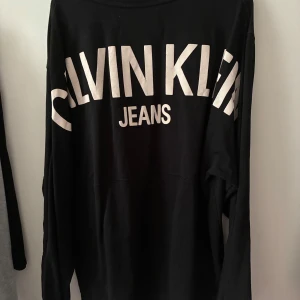 Svart långärmad tröja från Calvin Klein Jeans - Svart långärmad tröja från Calvin Klein Jeans med stort vitt tryck på ryggen och liten CK-logga framtill. Enkel och stilren design som passar perfekt till jeans eller joggers.