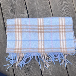 Ljusblå rutig halsduk från Burberry - Säljer en klassisk ljusblå halsduk från Burberrysi 100% lammull. Halsduken har ett rutigt mönster i beige, vitt och brunt samt fransar i ändarna. Perfekt accessoar för att lyfta din outfit. Några småhål och repor men annars bra skick och den är mjuk