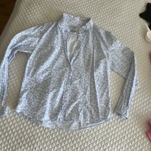 Blommig skjorta från Key West - Ljusblå och vit skjorta från Key West med blommigt mönster. Skjortan har långa ärmar, klassisk krage och knäppning framtill. Perfekt för en fräsch och avslappnad stil.