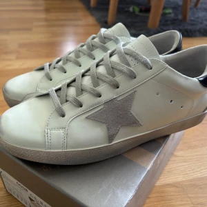 Vita sneakers från Golden Goose med grå stjärna - Säljer ett par vita sneakers från Golden Goose med grå mockadetaljer och en ikonisk stjärna på sidan. Skorna har svarta detaljer på hälen och grå snörning. Klassisk rund tå och platt sula. Perfekta för en trendig vardagslook.använda 2 gånger.Nypris runt 5000,mitt pris 2000!🙂