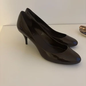 Bruna lackade pumps med klack - Säljer ett par bruna pumps i lackat material med rund tå och smal klack. Klassisk och stilren modell som passar perfekt till dig som gillar en elegant look. Skorna har en snygg glansig finish och är i storlek 40.