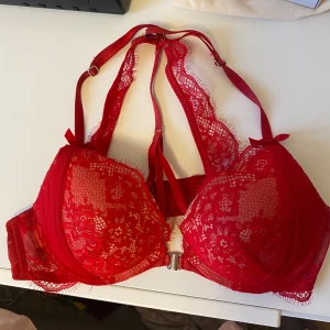 Röd spets-bh från Hunkemöller - Säljer en röd bh från Hunkemöller med vacker spets och dekorativ rosett framtill. Bh:n har justerbara axelband och snygga detaljer i metall framtill. Perfekt för dig som vill ha något extra fint under kläderna.