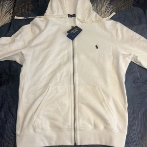 Vit hoodie från Polo Ralph Lauren - Snygg vit hoodie från Polo Ralph Lauren med dragkedja framtill och klassisk broderad logga på bröstet. Tröjan har huva med snörning och två stora fickor framtill. Perfekt för en chill och stilren look. 