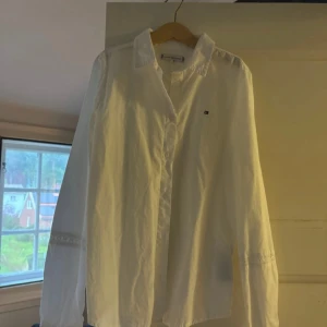 Vit skjorta från Tommy Hilfiger - Klassisk vit skjorta från Tommy Hilfiger med liten broderad logga på bröstet. Skjortan har lång ärm, knappar framtill och krage. Tillverkad i bomull och har en stilren, tidlös look som passar till många outfits.
