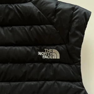 Svart dunväst från The North Face - Säljer en svart dunväst från The North Face med horisontella quiltade sömmar och hög krage. Västen har dragkedja framtill och broderad logga på bröstet. Perfekt för lager-på-lager och enkel att matcha med olika outfits.