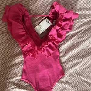 Rosa body med volang från Zara - Supersnygg rosa body från Zara med djup v-ringning och stora volanger. Bodyn har en tight passform och äben väldigt stretchigt i materialet. Perfekt för sommarens!