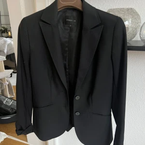 Svart kavaj från STOCKH LM, stl 32 - Klassisk svart kavaj från STOCKH LM i storlek 32. Kavajen har två knappar framtill, smala slag och fickor med lock. Materialet är en mix av ull, polyester och elastan för en bekväm passform. Perfekt för en stilren och tidlös look.