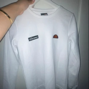 Vit långärmad tröja från Ellesse - Säljer en vit långärmad tröja från Ellesse med broderad logga och patch på bröstet. Tröjan har en sportig vibe med strukturerat tyg och rund hals. Perfekt för dig som gillar streetwear och enkel stil.
