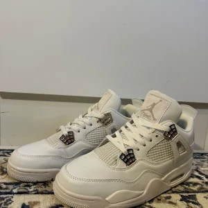 Nike Air Jordan 4 Retro Vit - Snygga Nike Air Jordan 4 Retro i helvitt skinn med silvriga detaljer och meshpaneler på sidorna. Klassisk Jumpman-logga på plösen och hälen. Skorna har snörning och en chunky sula som ger en sportig vibe. Perfekta för dig som gillar stilrena sneakers med ikonisk design.
