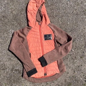 JH Collection brun och orange hoodiejacka - Snygg hoodiejacka från JH Collection i brun och orange med quiltad framsida, huva och dragkedja. Jackan har mjukt fleecetyg på insidan och är perfekt för lager-på-lager. Stora logga på bröstet och svarta muddar vid ärmslut. ( fina en matchande T-shirt) priset går att diskuteras.