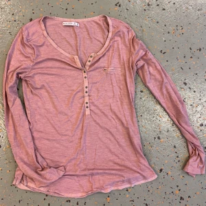 Ljusrosa långärmad tröja med knappar - Långärmad tröja i ljusrosa från pull&bear. Tröjan har en djup v-ringning med knappar framtill och en liten bröstficka. Mjuk och tunn i bomull, perfekt för lager-på-lager och chill dagar.