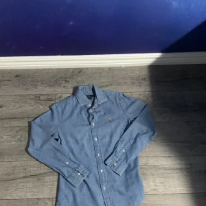 Blå skjorta från Polo Ralph Lauren - Snygg blå skjorta från Polo Ralph Lauren i custom fit-modell. Skjortan har klassisk krage, långa ärmar och knäppning framtill. Tillverkad i mjuk bomull och prydd med den ikoniska broderade loggan på bröstet. Perfekt för en clean och stilren look.