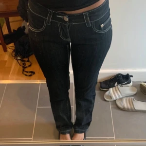 Mörkblåa jeans med vit kontrastsömmar - Säljer ett par mörkblåa jeans med vita kontrastsömmar och snygga silverfärgade nitar på bakfickorna. Modellen har låg midja och raka ben, klassisk femficksdesign och knappstängning framtill. Perfekta för dig som gillar en stilren men ändå cool look.