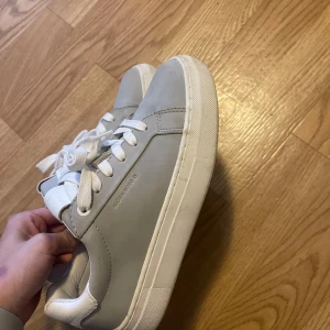 Gråa sneakers från Björn Borg, strl 37 - Snygga grå sneakers från Björn Borg med vita detaljer och snörning. Skorna har en klassisk rund tå, platt vit sula och är tillverkade i syntetmaterial. Perfekta för en clean och stilren look.
