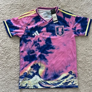Japan fotbollströja Adidas rosa/blå - Japan fotbollströja från Adidas i rosa och blått med coolt vågmotiv och JFA-emblem på bröstet. Tröjan har gula detaljer på axlar och ärmslut, samt klassisk Adidas-logga. Perfekt för dig som vill sticka ut på planen eller läktaren.