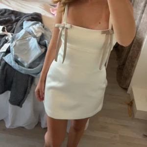 Vit kort klänning med rosetter från Zara - Supersöt vit kort klänning från Zara med raka axelband och fina rosetter framtill. Klänningen har en stilren, figurnära passform och är tillverkad i ett slätt, lätt glansigt material. Dragkedja i ryggen för enkel påklädning. Perfekt för sommarens alla tillfällen! Endast testad en gång! Prislapp kvar! Storlek S är slutsåld på hemsidan! Jag är 169 cm lång!