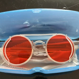 Runda glasögon med röda linser - Unika runda glasögon med transparent plastbåge och klarröda linser. Snygg detalj med metallfjädrar på sidorna som ger en futuristisk vibe. Perfekta för dig som vill sticka ut och addera färg till din stil.