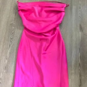 Rosa kort satinklänning med drapering - Snygg kort klänning i starkt rosa satin med draperad urringning och smala axelband. Klänningen har en figurnära passform och en slät baksida med dragkedja. Perfekt för dig som vill sticka ut med en färgstark look.