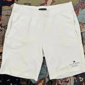 Vita shorts från Tommy Hilfiger - Snygga vita shorts från Tommy Hilfiger med bred ribbad midja och diskret logga framtill. Mjukt bomullsmaterial, två sidofickor och en bakficka med liten flagga. Perfekta för sommardagar och chill vibes.