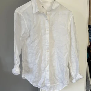 Vit linneskjorta från Uniqlo - Klassisk vit skjorta i linne från Uniqlo med lång ärm och knappar framtill. Skjortan har en avslappnad passform och mjuk känsla, perfekt för en stilren och enkel look. Snygg att bära både knäppt och öppen över t-shirt.