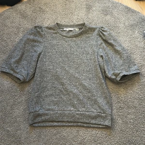 Grå tröja med puffärm från H&M - Säljer en grå tröja från H&M med puffiga korta ärmar och rund halsringning. Tröjan är i mjukt material och har en enkel, stilren look som passar perfekt till jeans eller kjol. Perfekt för dig som gillar en chill men ändå trendig stil.