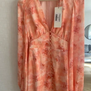 Adore kort blommig klänning i Peach - Superfin kort klänning från Adore i en peachy orange nyans med blommigt mönster. Klänningen har v-ringning, långa puffärmar och knappar framtill. Materialet känns lätt och flowy, perfekt för sommarens alla tillfällen. Sitter snyggt med markerad midja och ballongärmar. Oanvända i förpackning säljer flera storlekar , 34,36,40,48
