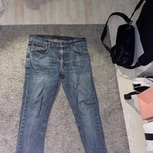 Nudie Jeans blå slim fit jeans - Säljer ett par klassiska blå jeans från Nudie Jeans med snygg tvätt och kontrastsömmar. Modellen är slim fit med raka ben och fem fickor. Jeansen har detaljerade sömmar på bakfickorna och är tillverkade i bomull. Perfekta för en avslappnad och stilren look.