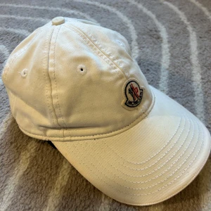 Vit keps från Moncler - Snygg vit keps från Moncler med klassisk böjd skärm och broderad logga framtill. Kepsen är tillverkad i bomull och har ventilerande hål upptill för extra komfort. Perfekt accessoar för en clean och sportig look.
