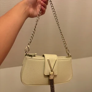 Beige axelväska från Valentino - Snygg beige axelväska från Valentino med kedjerem i metall och V-logga framtill. Väskan är i skinnimitation med struktur och har dekorativt hänge i metallkedja. Perfekt storlek för det viktigaste och ger en lyxig känsla till din outfit. Enda defekten är att det som hämger från v:et har rostat. Nypris 1200kr!!! 