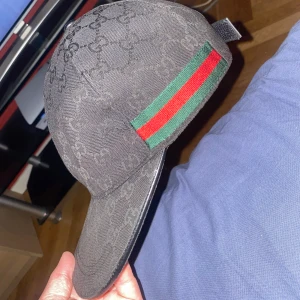 Svart keps från Gucci med GG-mönster - Snygg svart keps från Gucci med klassiskt GG-monogram över hela kepsen och ikoniska gröna och röda ränder på sidorna. Justerbar baktill och tillverkad i slitstarkt material. Perfekt för dig som vill ha en stilren och lyxig accessoar.