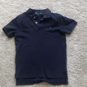 Marinblå pikétröja från Polo Ralph Lauren - Snygg marinblå pikétröja från Polo Ralph Lauren med klassisk krage och två knappar framtill. Tröjan har korta ärmar och den ikoniska röda broderade loggan på bröstet. Tillverkad i 100% bomull för en skön och avslappnad känsla.
