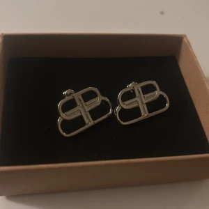Balenciaga BB örhängen i silver - Snygga örhängen från Balenciaga i silverfärgad metall med den ikoniska BB-loggan. De har en stilren och modern design som verkligen sticker ut. Perfekta för dig som vill ha något exklusivt och trendigt i smyckesamlingen.