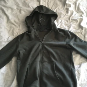 Mörkgrön hoodiejacka Peak Performance - Mörkgrön hoodiejacka från Peak Performance med dragkedja framtill och två fickor. Klassisk stil med broderad logga på bröstet och mjukt material som känns skönt mot huden. Perfekt för dig som gillar en clean och sportig look.