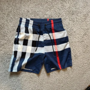 Burberry shorts - Snygga badshorts från Burberry med klassiskt rutigt mönster i blått, vitt, svart och en röd rand. Shortsen har snörning i midjan och är tillverkade i ett lätt syntetmaterial som torkar snabbt. Perfekta för stranden eller poolen i sommar. Självklart äkta 