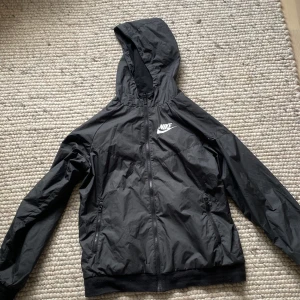 Svart Nike vindjacka med huva - Svart vindjacka från Nike med huva och dragkedja hela vägen. Jackan har två fickor med dragkedja och en vit Nike-logga på bröstet. Lätt och smidig modell i syntetmaterial, perfekt för blåsiga dagar.