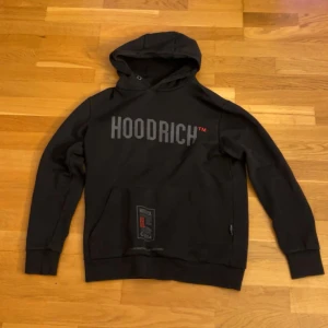 Svart hoodie från Hoodrich med tryck - Svart hoodie från Hoodrich med stor logga framtill och coolt tryck på ryggen. Tröjan har huva, känguruficka och långa ärmar. Snygga röda och grå detaljer på både fram- och baksida samt på ena ärmen. Perfekt för dig som gillar streetwear.
