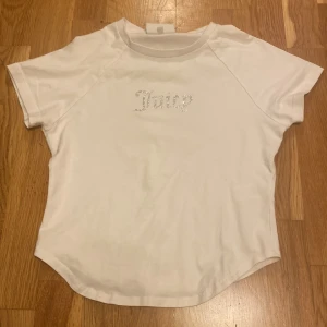 Vit Juicy Couture t-shirt med strass - Vit t-shirt från Juicy Couture med korta ärmar och rund hals. Framsidan har texten 'Juicy' i glittrande strass. T-shirten är i mjuk bomull och har en klassisk, lite feminin passform. Perfekt för dig som gillar en clean men ändå glam look🤍