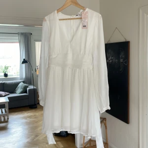 Klänning - Superfin vit kortklänning med v-ringning och långa, transparenta ärmar. Klänningen har ett markerat midjeband och lätt utsvängd kjol i tunt, flowy material. Perfekt för sommarens alla tillfällen och ger en fin look. Klänningen är helt ny och kostar 750kr mitt pris är 200kr😀