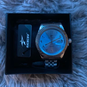 Silverfärgad klocka från RICECGO - Stilren klocka från RICECGO i silverfärg med arabiska siffror på urtavlan. Klockan har en rund boett, räfflad kant och länkarmband i metall. Urtavlan är ljus med datumvisning vid klockan tre. Perfekt accessoar för dig som gillar klassisk design.