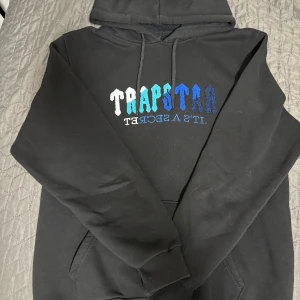 Trapstar Hoodie - Svart Trapstar Hoodie i storlek XXL men kan passa L. Framsidan klassisk Trapstar-logga i vitt, turkos och blått samt texten 'It's a secret' i blått. Luva med snörning och känguruficka. Bredd: 57 cm och längd:70 cm