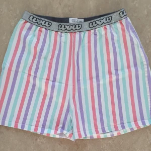 Shorts - Färgglada shorts från Wow med vertikala ränder i lila, rosa, turkos och vitt. De har en bred, glittrig silverfärgad resår i midjan med Wow-logga. Perfekta för sommarens chill dagar och riktigt sköna att ha på sig.