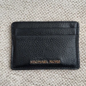 Svart korthållare Michael Kors skinn - Stilren svart korthållare från Michael Kors i äkta skinn med guldfärgad logga framtill. Plats för flera kort på både fram- och baksidan.  
