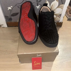 Exklusiva Christian Louboutin Sneakers med Signaturröd Sula - Stilrena Christian Louboutin sneakers i svart mocka med ikoniska röda sulor och nitar framtill för en exklusiv, iögonfallande look. Äkta vara – levereras med originalkartong och äkthetskort. Perfekt för dig som vill sticka ut med klass pris kan diskuteras