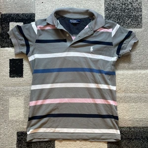 Randig pikétröja från Ralph Lauren - Snygg pikétröja från Ralph Lauren i grått med breda ränder i blått, vitt, rosa och marinblått. Klassisk krage, korta ärmar och broderad logga på bröstet. Perfekt för en chill och stilren look.