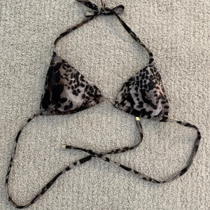 Leopard bikiniöverdel Calzedonia - Snygg trekantsbikini från Calzedonia med leopardmönster i svart och bruna toner. Bikinin har knytband i nacken och ryggen, samt små guldfärgade detaljer på snörena. Perfekt för stranden eller poolen i sommar! As snygg nypris ca 350 kr