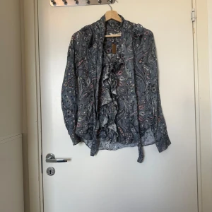 Mönstrad blus med volang och knyt - Supersnygg blus i en mörkblå ton med färgglada detaljer och blommigt mönster. Långärmad modell med volang framtill, v-ringning och knytband vid halsen. Tunt, glansigt material som känns lyxigt och skönt mot huden. Från märket Odd Molly i storlek 1 som ungefär motsvarar en storlek S. Mycket fint skick, jag hittar inga synlig anmärkningar eller defekter.  