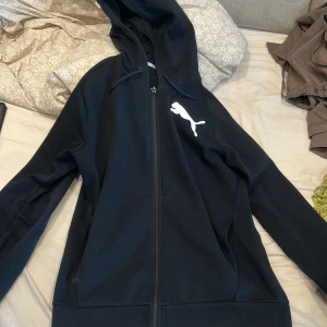Svart hoodiejacka från Puma - Svart hoodiejacka från Puma med dragkedja och stor vit logga på bröstet. Jackan har huva med snörning och meshfoder på insidan. Perfekt för chill dagar eller när du vill ha en sportig vibe.